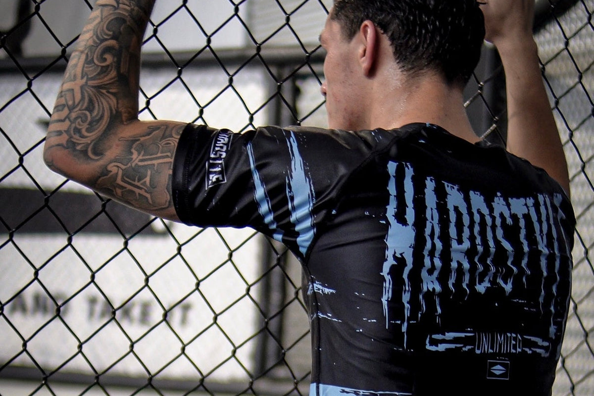 A man seen from behind wearing a black and light blue rash guard with a distressed 'HANDS ON THE FENCE' graphic and matching light blue training shorts, standing inside an MMA cage.